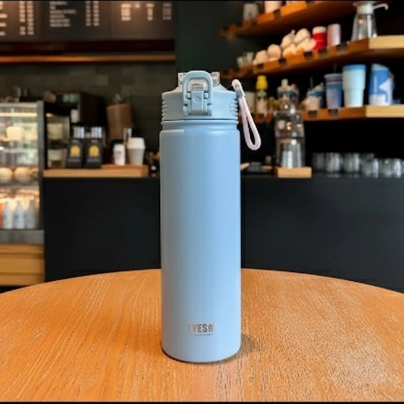 Bouteille isotherme en inox bleu TYESO posée sur une table de café, avec bouchon sport anti-fuite et dragonne, design moderne, idéale pour garder les boissons chaudes ou froides plusieurs heures.