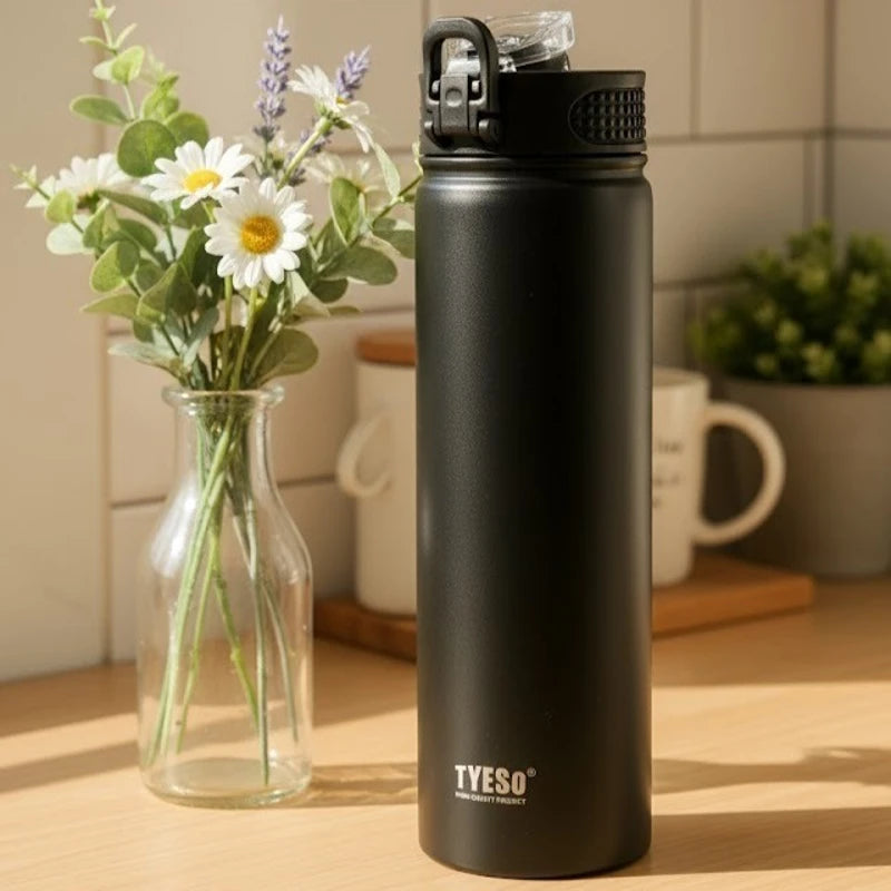 Bouteille isotherme en inox noir mat TYESO posée sur une table de cuisine, entourée de tasses et de fleurs, idéale pour garder les boissons chaudes ou froides tout au long de la journée.