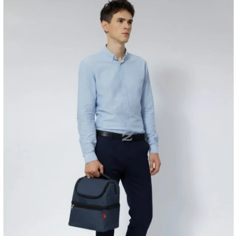 Homme debout tenant un sac isotherme bleu par la poignée, habillé en chemise claire et pantalon foncé.