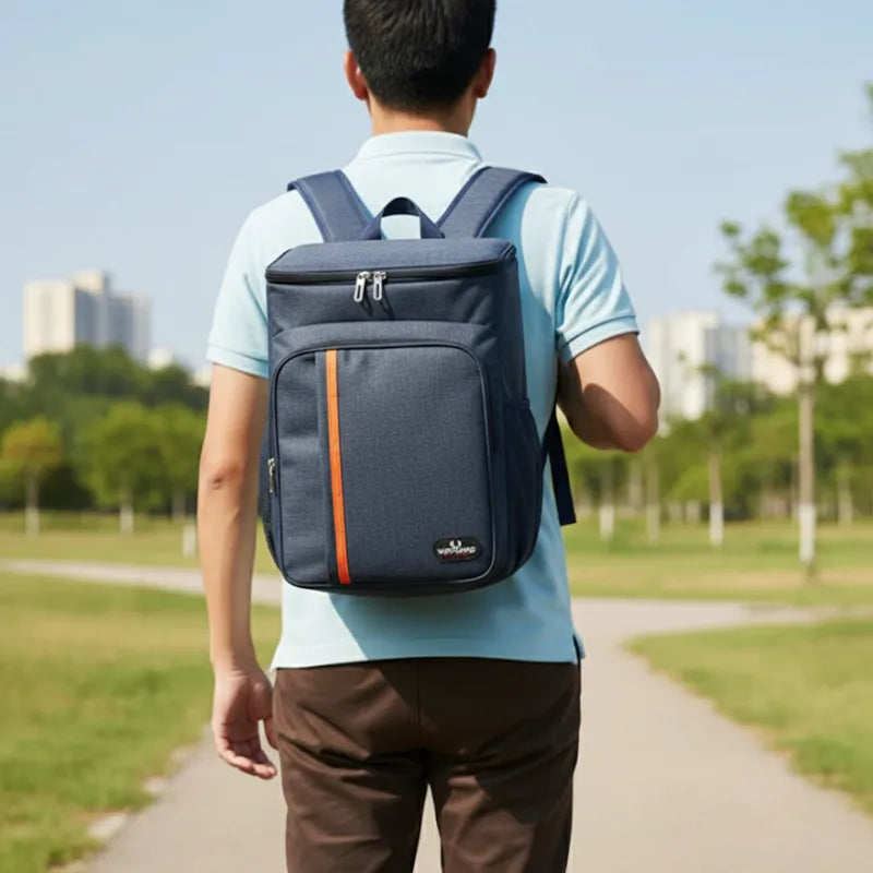 Homme de dos marchant dans un parc avec un sac à dos isotherme bleu marine muni d’une bande orange, en extérieur par temps ensoleillé.