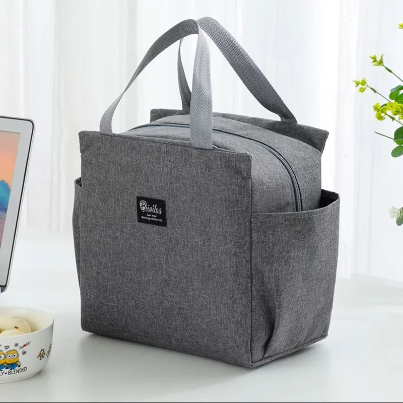 Sac isotherme repas gris à poignées avec fermeture zippée, format compact avec poches latérales, posé sur une table, conçu pour transporter un déjeuner ou un repas du midi.