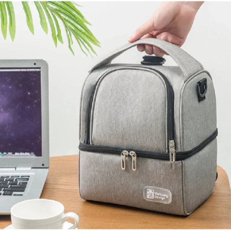 Sac isotherme bébé gris tenu par la poignée, posé sur un bureau près d’un ordinateur et d’une tasse.