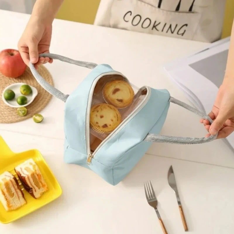 Femme en tablier tenant les poignées d’un sac isotherme bleu pastel ouvert vu de dessus, laissant apparaître des pâtisseries à l’intérieur. Le sac est posé sur une table avec un sandwich, des fruits et des couverts, illustrant la préparation d’un repas maison à emporter.