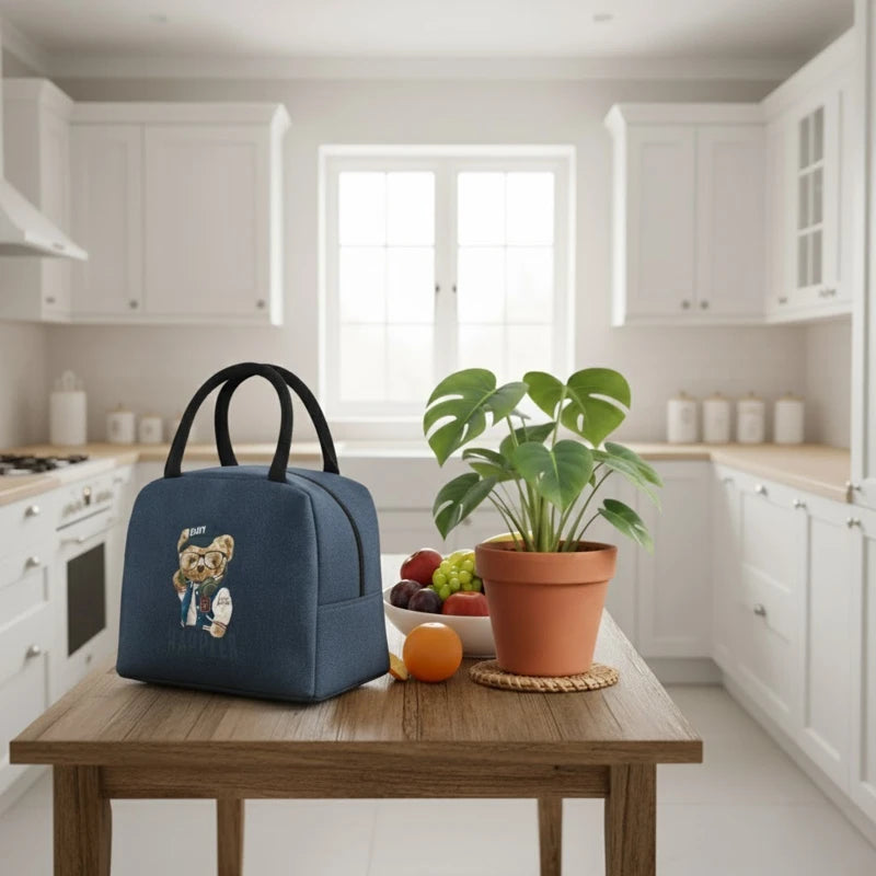 Sac isotherme enfant bleu avec motif ourson Happier posé sur une table en bois dans une cuisine lumineuse, à côté d’un bol de fruits et d’une plante verte.