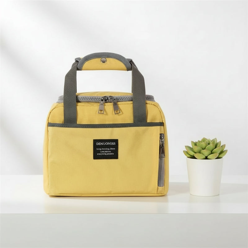 Sac isotherme jaune posé sur une table blanche, avec anses grises et poche frontale zippée, présenté dans un style minimaliste avec une plante décorative.