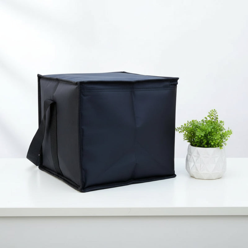 Sac isotherme noir de 50 L en forme de cube, posé sur une table blanche, avec poignée latérale et plante décorative à côté.