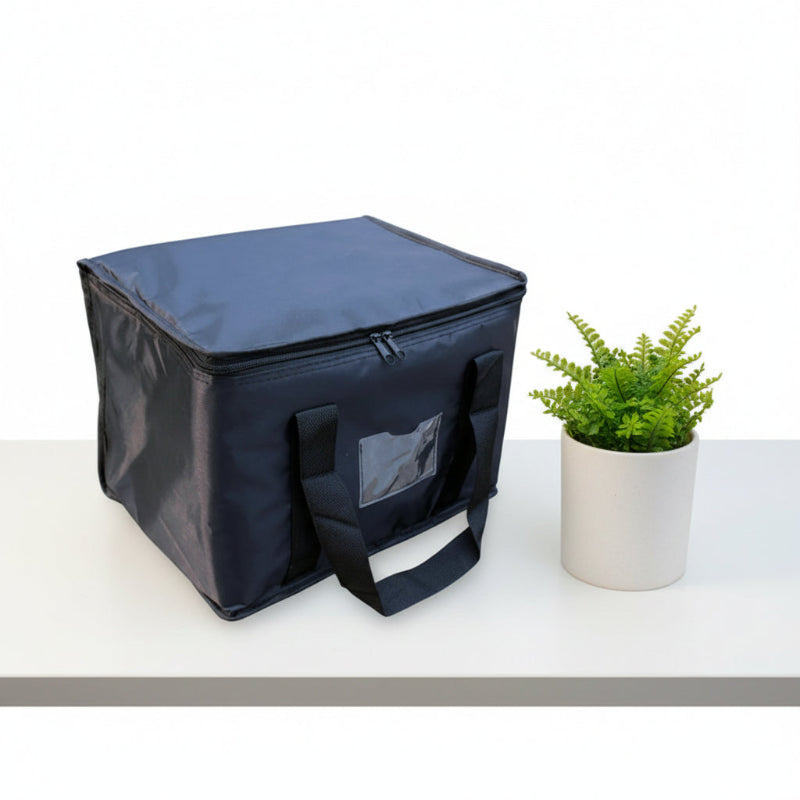 Sac isotherme bleu marine rectangulaire posé sur une table blanche, avec poignées noires et petite poche transparente sur le devant, accompagné d’une plante en pot.