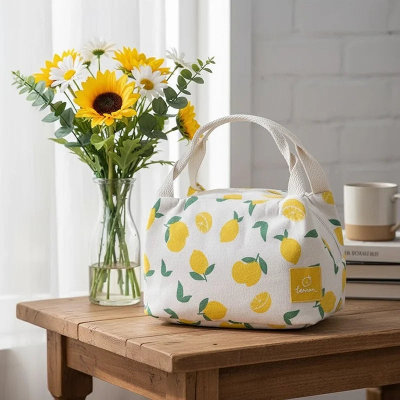 Sac isotherme à motif citrons posé sur une table en bois près d’un vase de tournesols et de marguerites, dans une pièce lumineuse.
