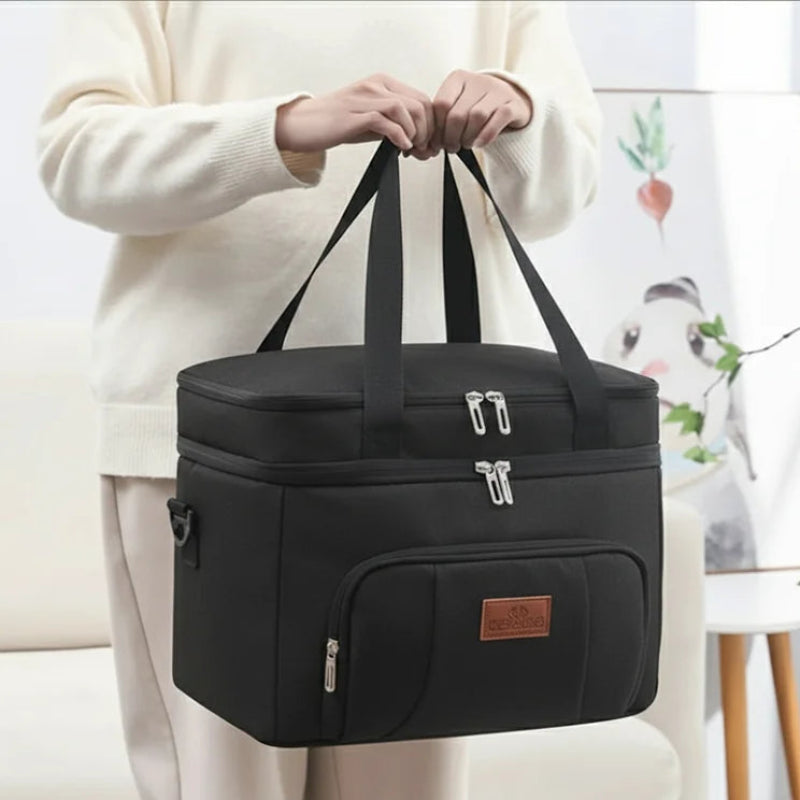 Sac isotherme repas noir porté à la main par une personne en intérieur, avec poignées noires et grande poche frontale.