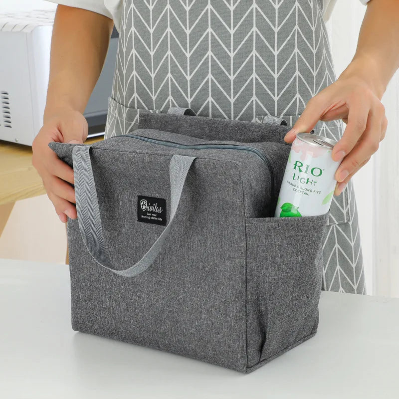 Sac isotherme repas gris avec poignées et fermeture zippée, posé sur une table, doté de poches latérales, utilisé pour transporter un déjeuner avec boisson et repas du midi.
