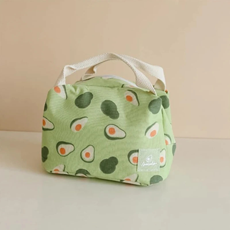 Sac isotherme vert à motifs d’avocats, posé sur une surface beige, avec des anses en tissu blanc et un fond uni clair en arrière-plan.