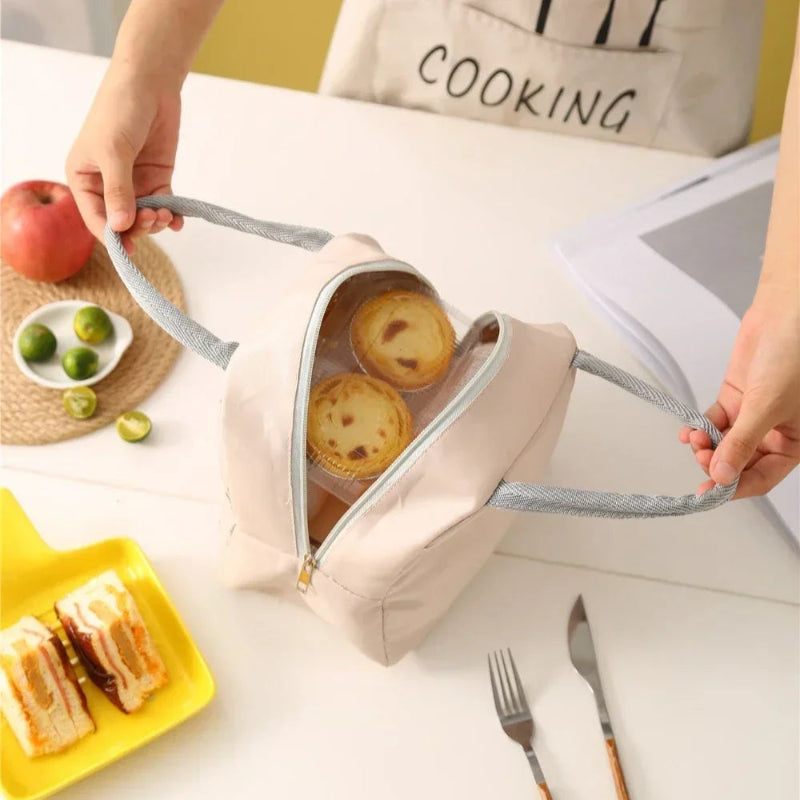 Sac isotherme repas femme beige sans motif, vu de dessus, ouvert, contenant deux tartelettes dans un emballage transparent. Il est tenu par les poignées grises, posé sur une table avec des couverts et des assiettes.
