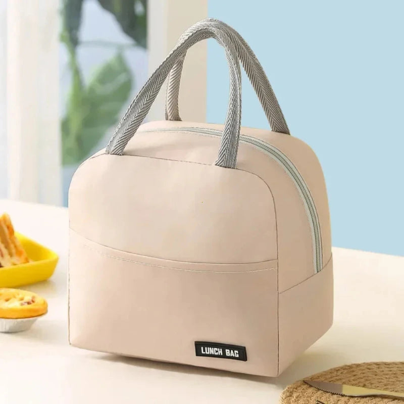Sac isotherme repas femme beige sans motif, vu de trois quarts, posé sur une table claire avec des poignées grises et une fermeture éclair claire, dans un décor lumineux.
