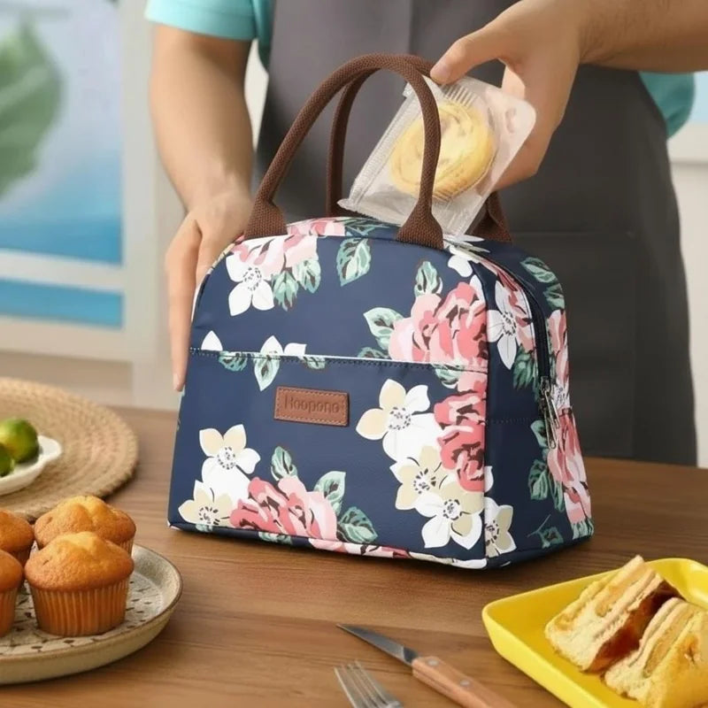 main plaçant une pâtisserie dans un sac isotherme repas bleu marine à motifs floraux, posé sur une table en bois avec muffins et tranches de gâteau dans une assiette jaune