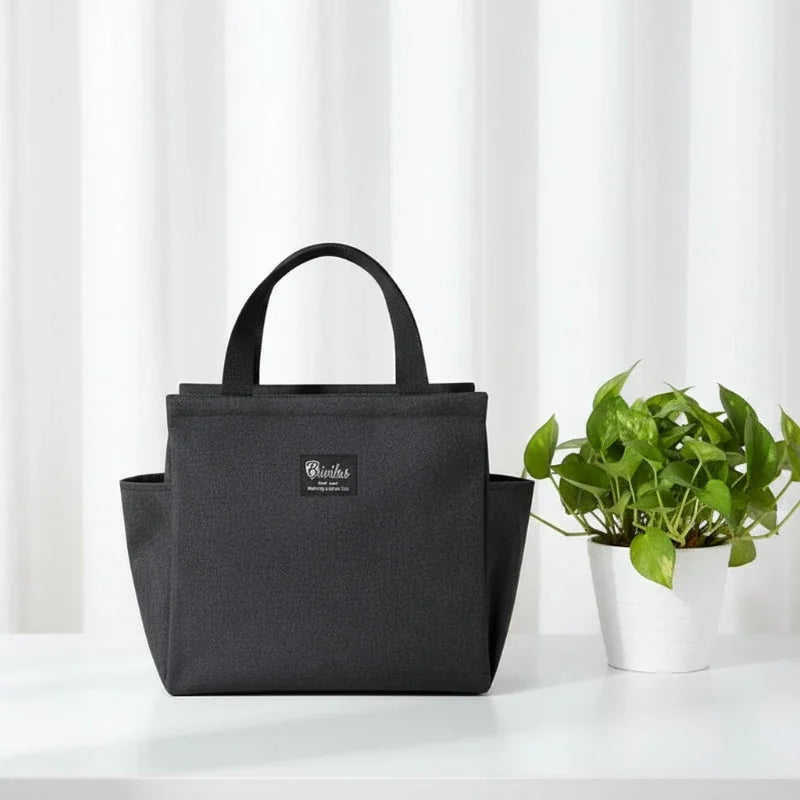Sac isotherme repas noir anthracite type lunch bag posé sur une table, avec plante verte à côté, idéal pour transporter un déjeuner au bureau.