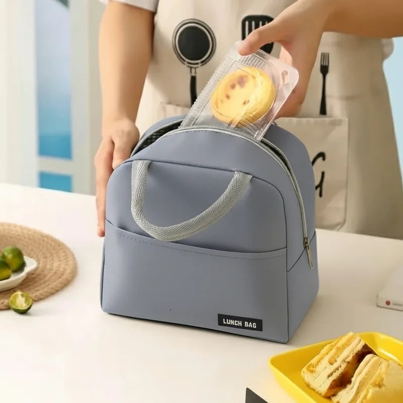 Personne rangeant une pâtisserie dans un sac isotherme repas gris sur une table de cuisine, avec une assiette de sandwich et des citrons verts en arrière-plan.