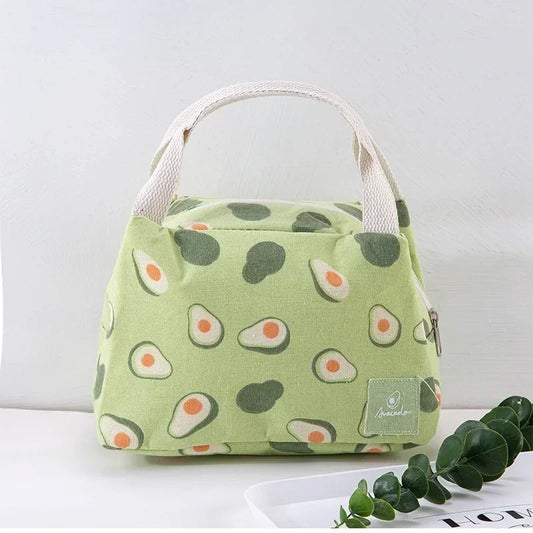 Sac isotherme vert clair à motifs d’avocats, avec des anses beiges, posé sur une table blanche à côté de feuilles vertes décoratives.