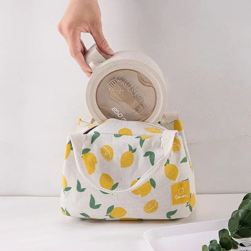 Main plaçant une lunch box beige dans un sac isotherme repas motif citron jaune, avec anses en coton tressé et fond blanc minimaliste.