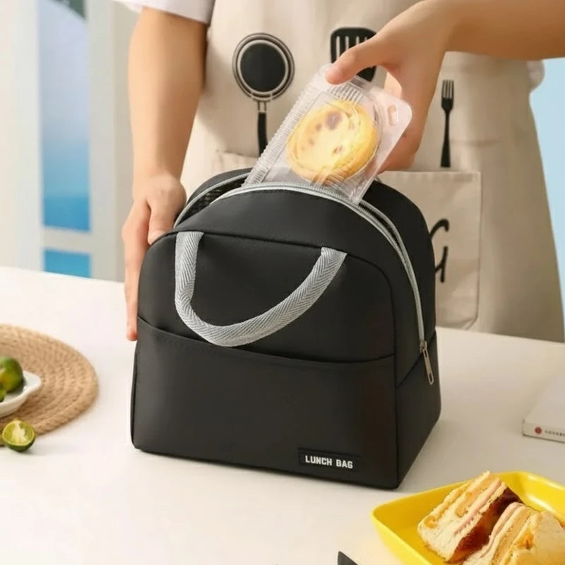 Personne en tablier beige plaçant un petit gâteau dans un sac isotherme noir posé sur une table blanche, à côté d’une assiette jaune contenant un sandwich coupé en deux.