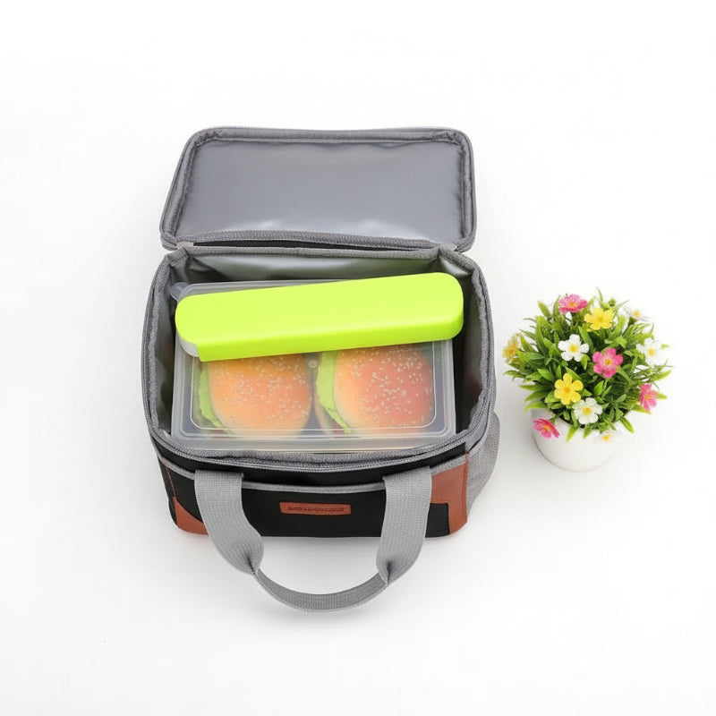 Sac isotherme repas noir et gris ouvert avec une boîte de déjeuner et un pain de glace, posé sur une table blanche avec une plante fleurie.