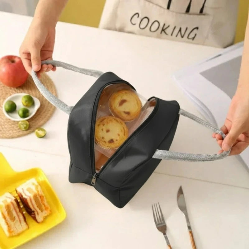 Sac isotherme noir ouvert vu du dessus, tenu par les deux poignées grises, contenant deux petits gâteaux dans une boîte transparente, posé sur une table blanche avec une assiette jaune et des sandwichs à côté.