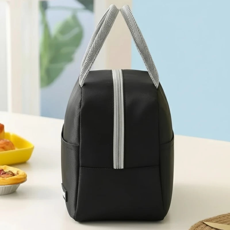 Sac isotherme repas noir vu de profil, posé sur une table blanche, avec poignées grises dressées et fermeture éclair argentée, en arrière-plan un mur bleu clair et une feuille verte floue.