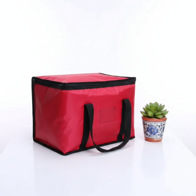 Sac isotherme rouge pour transport de repas, vu en angle, avec poignées noires et plante décorative sur table blanche.