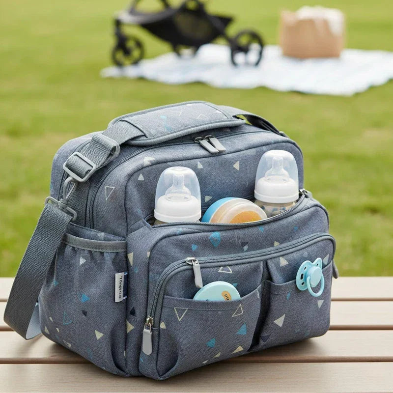 Sac isotherme pour bébé avec poches avant contenant des biberons et accessoires, posé sur une table en extérieur.