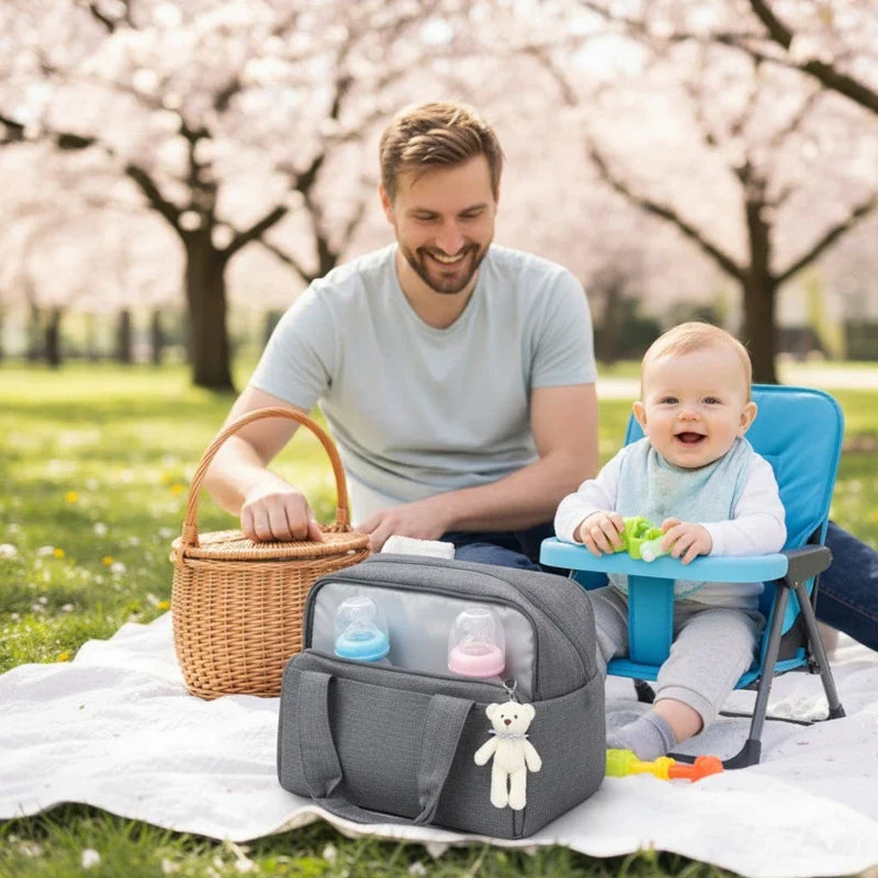 Scène familiale en plein air montrant un père souriant à côté de son bébé assis sur une chaise de pique-nique, avec un sac isotherme gris contenant des biberons et un doudou, sur une couverture étendue dans un parc fleuri.