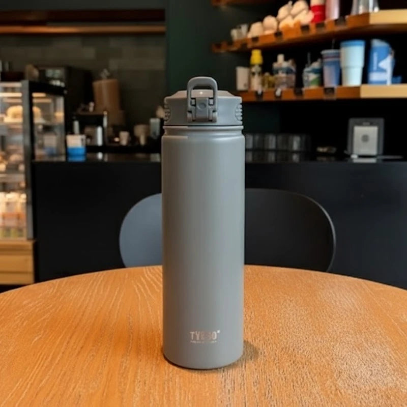 Bouteille isotherme en inox gris TYESO posée sur une table de café, bouchon sport anti-fuite, design sobre et moderne, idéale pour garder les boissons chaudes ou froides plusieurs heures.