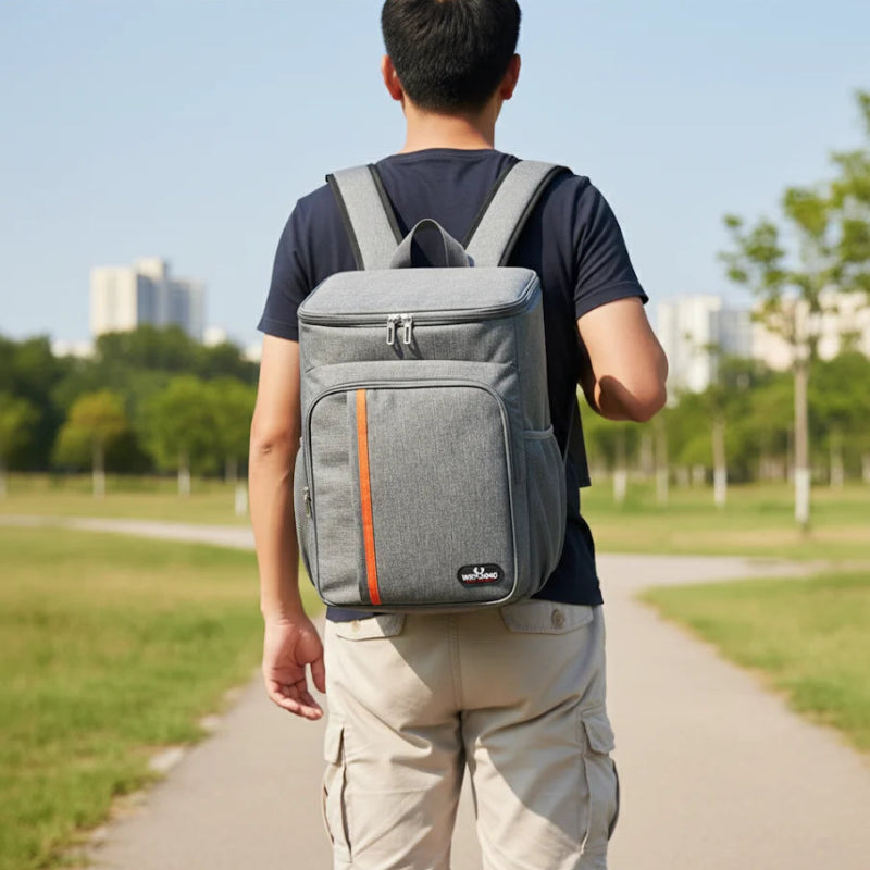 Homme portant un sac à dos isotherme gris avec bande orange, marchant sur un chemin en plein air dans un parc, idéal pour les pique-niques ou les déplacements quotidiens.