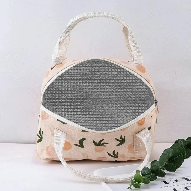Sac isotherme repas beige à motifs pêches et feuilles vertes, posé sur une table en bois dans une cuisine lumineuse avec tasse et planches à découper en arrière-plan.