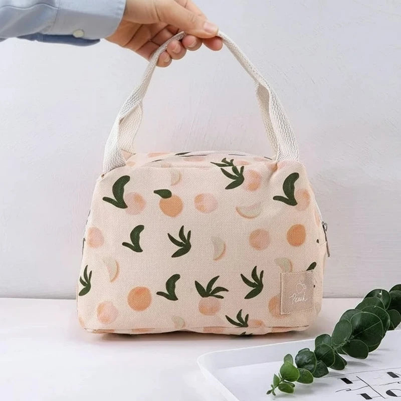 Sac isotherme beige à motifs pêches et feuilles vertes, tenu par une main, avec anses en tissu blanc, idéal pour transporter un repas au travail ou en pique-nique.
