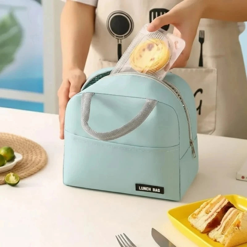 Femme en tablier plaçant un gâteau dans un sac isotherme bleu pastel ouvert, posé sur une table avec un sandwich et des fruits. Le sac en tissu Oxford, muni de poignées grises et d’une fermeture éclair argentée, illustre une scène de préparation de repas à emporter.