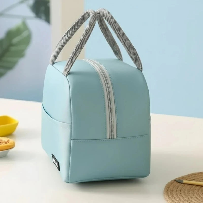 Sac isotherme bleu pastel vu de côté, posé sur une table blanche. On distingue ses poignées grises croisées et sa fermeture éclair argentée, dans une scène lumineuse avec pâtisseries et fond bleu clair, évoquant un déjeuner prêt à emporter.
