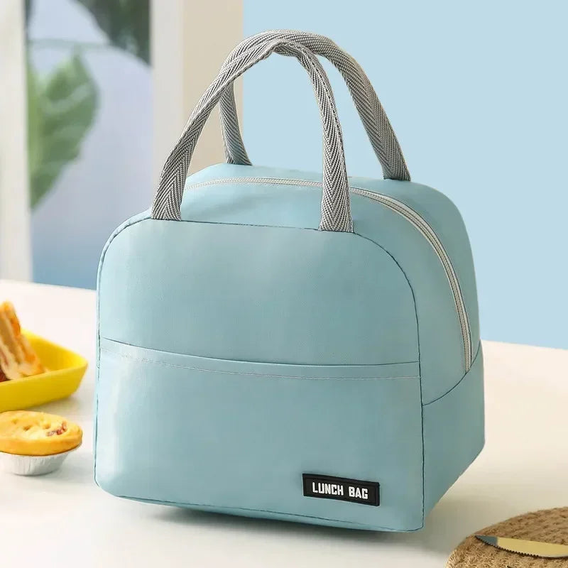 Sac isotherme bleu pastel vu de face, posé sur une table blanche avec poignées grises et fermeture éclair argentée. En arrière-plan, une assiette de pâtisseries et un décor lumineux bleu clair créent une ambiance douce et propre, idéale pour illustrer un déjeuner à emporter.