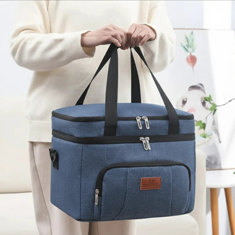 Sac isotherme repas bleu porté à la main par une personne en intérieur, avec poignées noires et grande poche frontale.