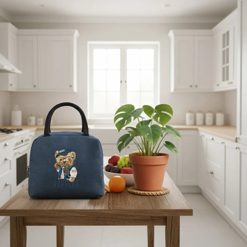 Sac isotherme enfant bleu avec motif ourson “Happier”, posé sur une table dans une cuisine lumineuse avec un plant et des fruits à côté.