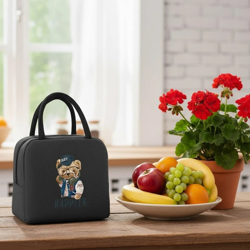 Sac isotherme enfant noir avec motif ourson, posé sur une table de cuisine à côté d’un bol de fruits, dans une ambiance lumineuse et naturelle.