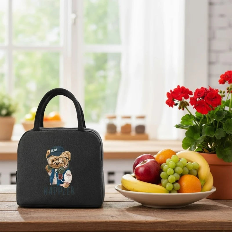 Sac isotherme pour enfant de couleur noire avec motif ours, posé sur une table de cuisine à côté d’un plat de fruits frais et d’une plante fleurie.