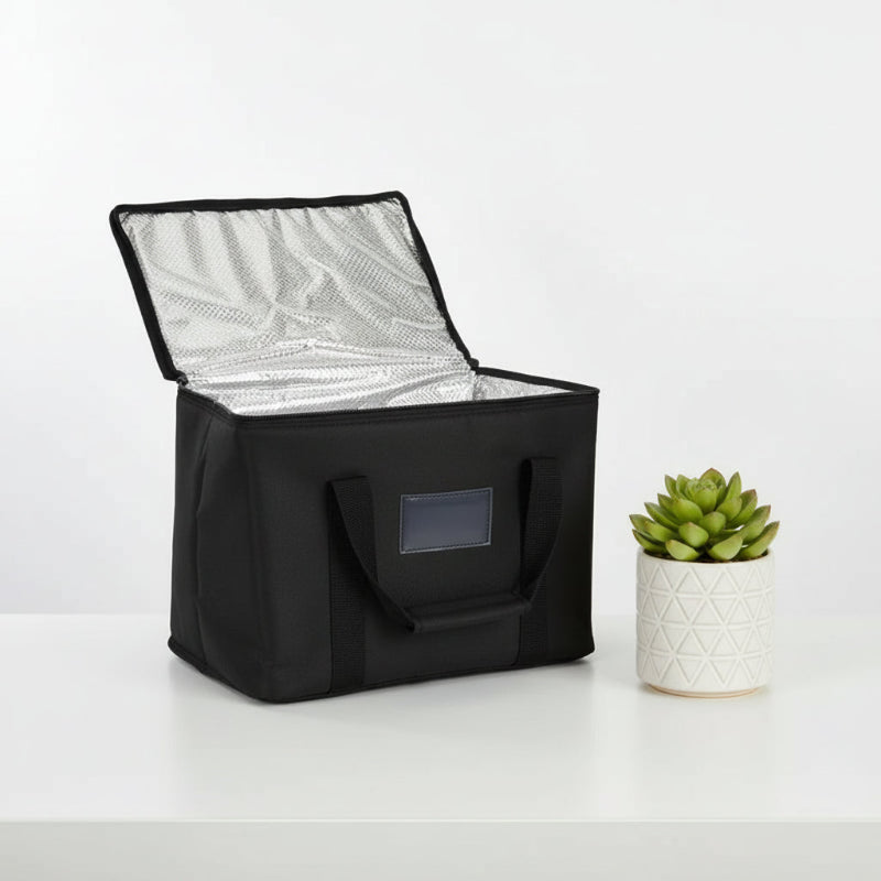 Sac isotherme de livraison noir 50 L ouvert, avec intérieur aluminium visible, posé sur une table blanche à côté d’une plante décorative.