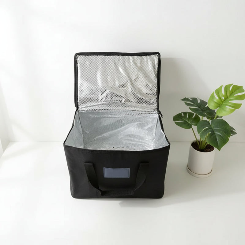 Sac isotherme noir ouvert montrant l’intérieur aluminium thermique, posé sur une table blanche avec une plante décorative.