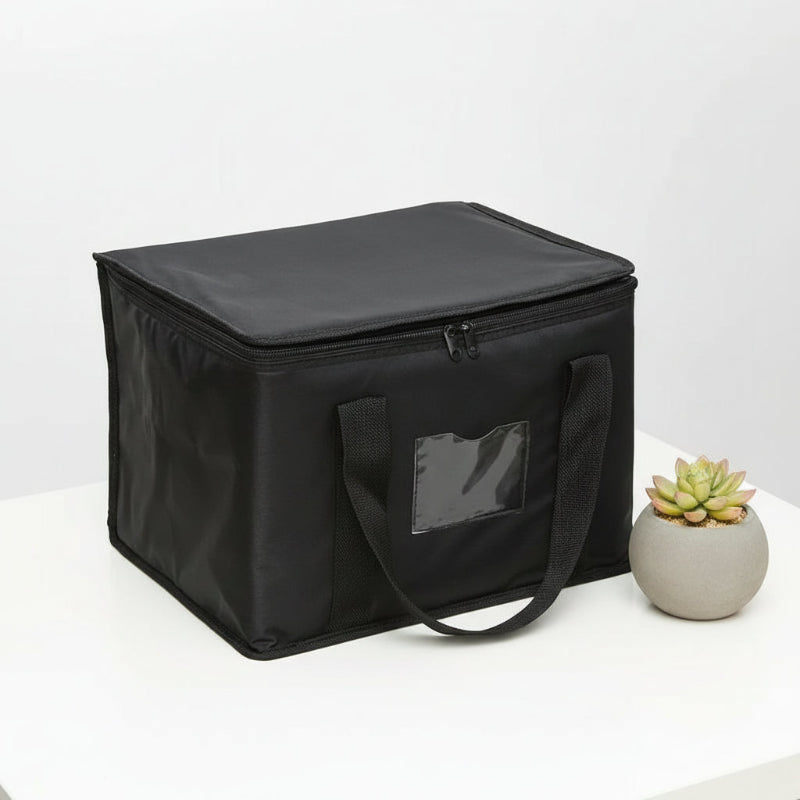 Sac isotherme noir rectangulaire posé sur une table blanche, avec poignées en tissu et petite plante décorative à côté.