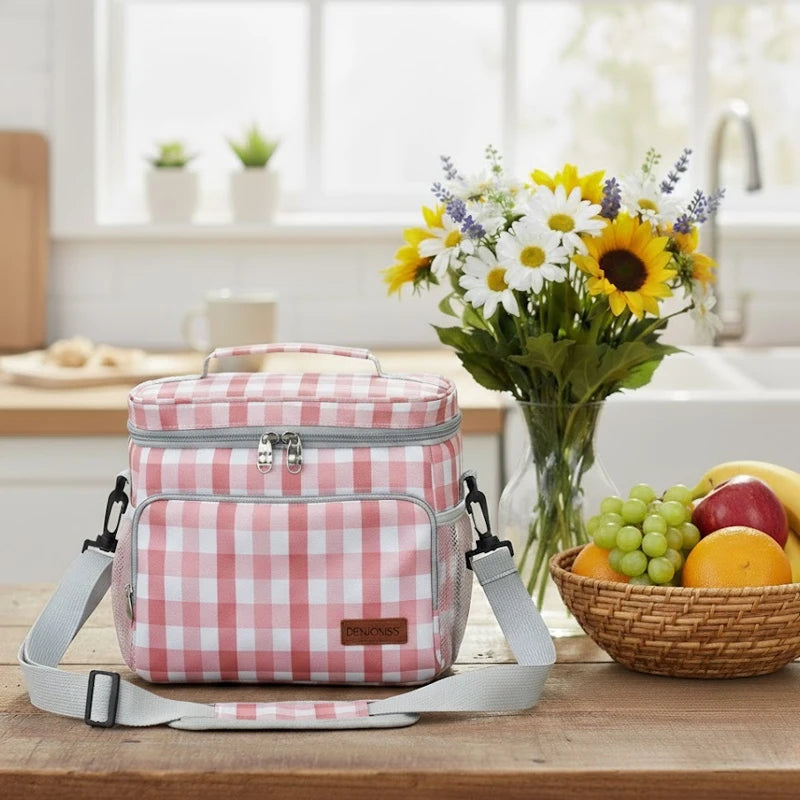 Sac isotherme motif vichy rose posé sur une table en bois dans une cuisine lumineuse, avec bandoulière grise, bouquet de fleurs et panier de fruits en arrière-plan