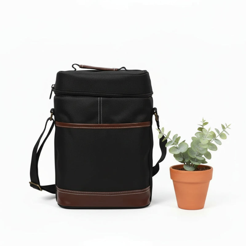 Sac isotherme noir pour bouteille avec finitions cuir marron et bandoulière, posé sur une table blanche à côté d’une plante.