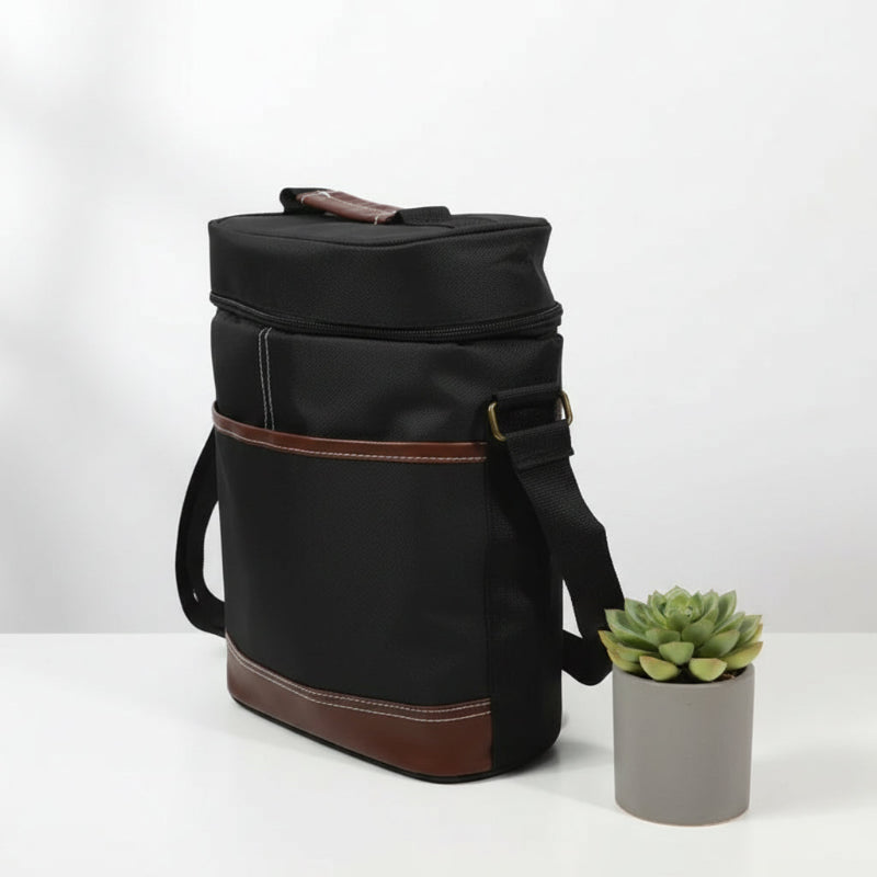 Sac isotherme noir pour bouteille avec finitions cuir marron vu de côté, posé sur une table blanche à côté d’une plante succulente.