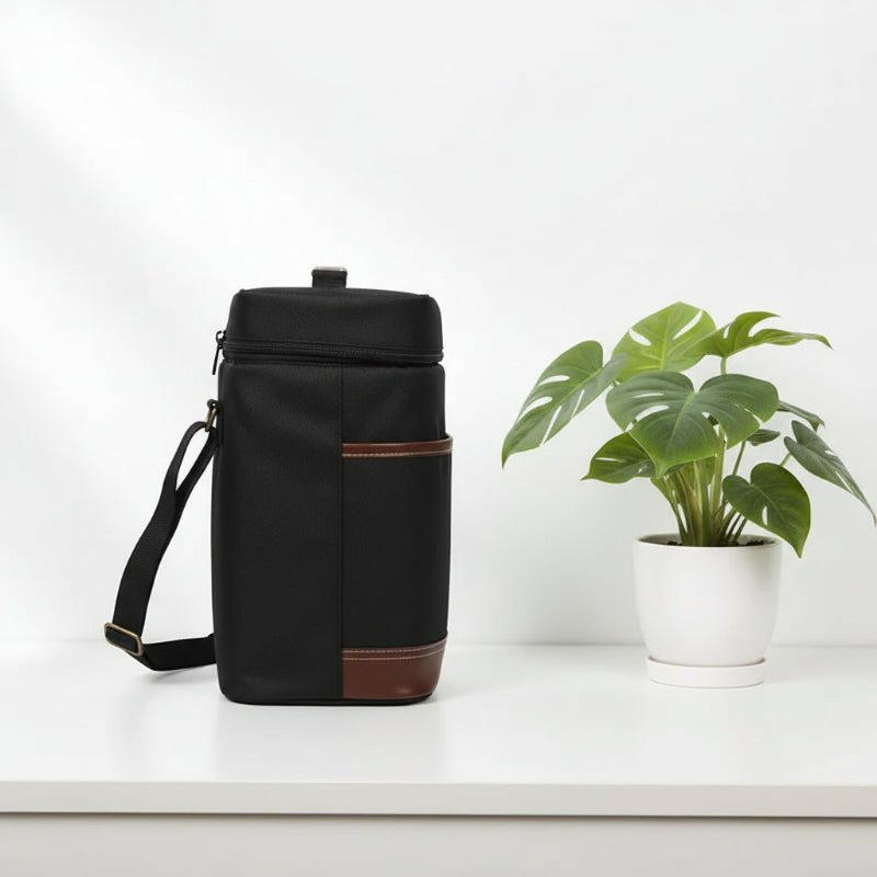 Sac isotherme noir pour bouteille avec finitions cuir marron vu de profil, posé sur une table blanche à côté d’une plante verte.
