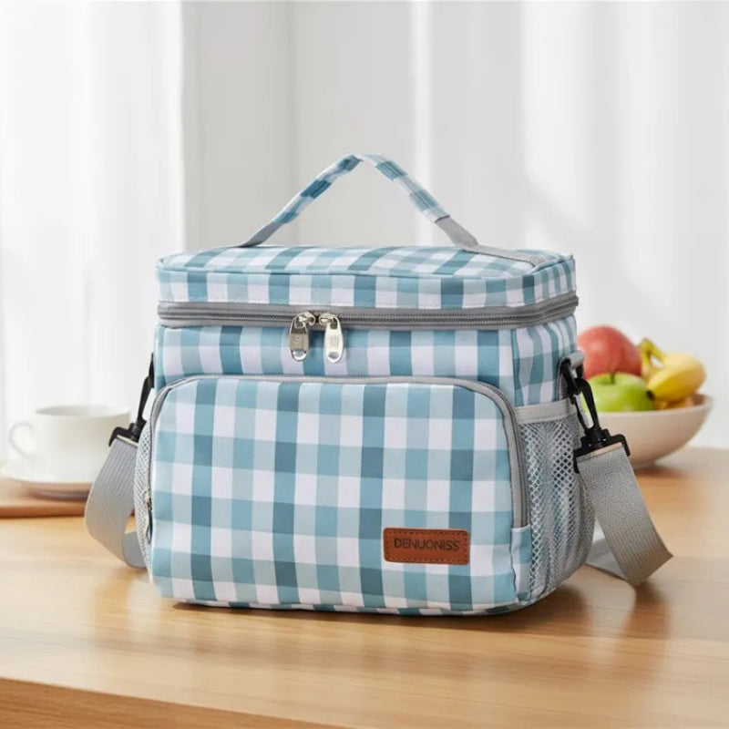 Sac isotherme repas à carreaux vert et blanc posé sur une table de cuisine, avec une tasse de café et un bol de fruits en arrière-plan.