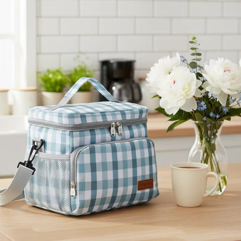 Sac isotherme repas à carreaux vert et blanc posé sur une table de cuisine, à côté d’un bouquet de fleurs et d’une tasse de café.