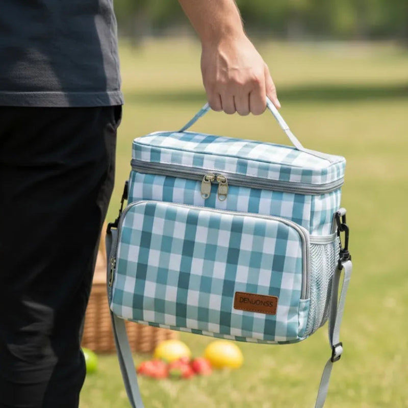 Sac isotherme repas à carreaux vert et blanc porté à la main en extérieur, avec un panier de pique-nique et des fruits en arrière-plan.